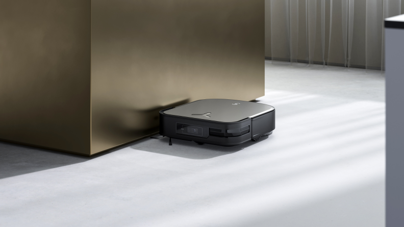 A square, black robot vacuum sits on a gray floor, flush against a gold wall.