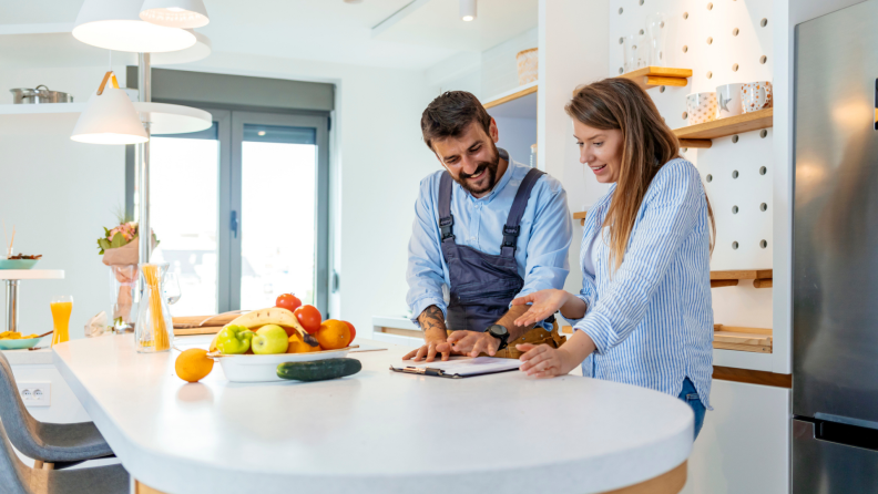 Woman and man smiling while standing together in kitchen while both looking at a clipboard on the counter.