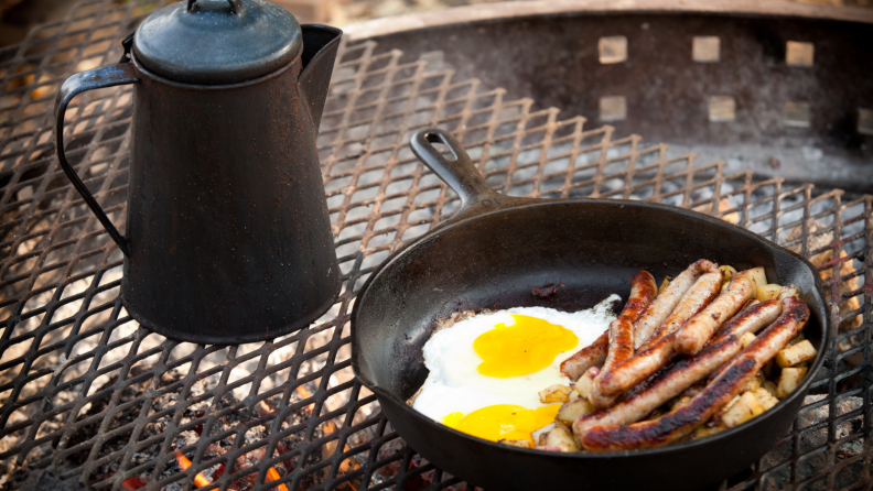 In the foreground, there's a cast-iron skillet with two eggs and some bacon in it. To the left of the skillet, there's a cast-iron kettle. Both are on a makeshift outdoor stove.