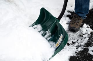 A close-up of a snow shovel digging into a snow bank. The person holding it is attempting to clear a walkway that's covered in a few inches of snow. .