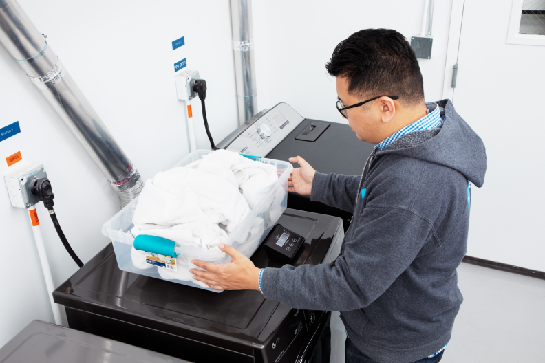 Person placing clear storage bin filled with white shirts on top of scale resting on dryer inside of white room.