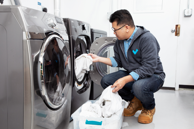 Person crouched in front dryer while inserting white towels into drum inside of white room.