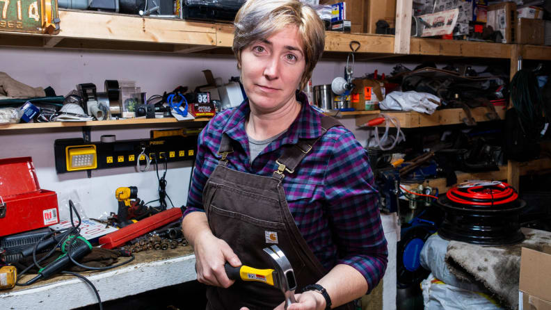 The author stands by her busy workbench, in a work apron, hammer in hand.