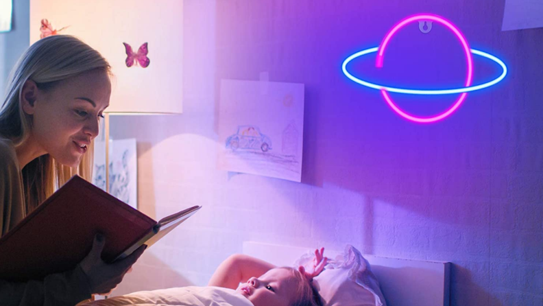 Mother reading a book to a child in bed with a pink and blue neon Saturn-inspired sign on the wall