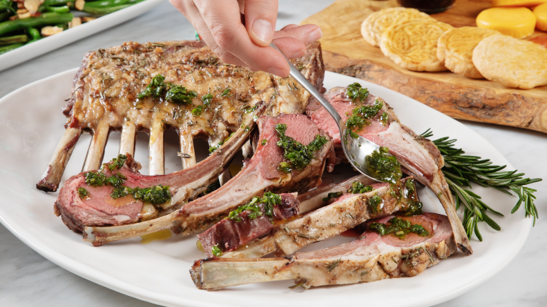 Rack of lamb served on a white platter with a person drizzling herb sauce over it.
