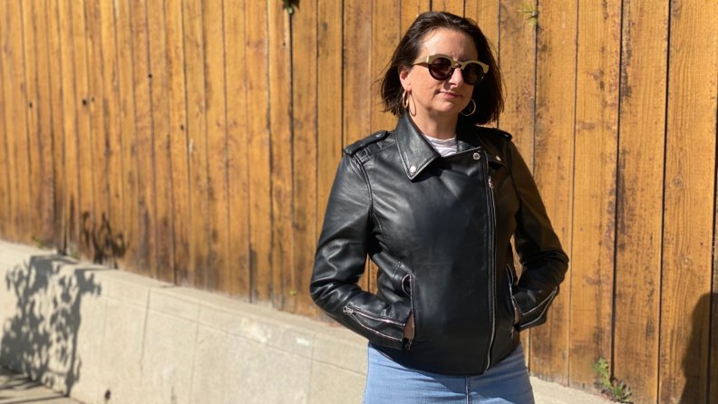 A woman stands against a fence in a black leather jacket.