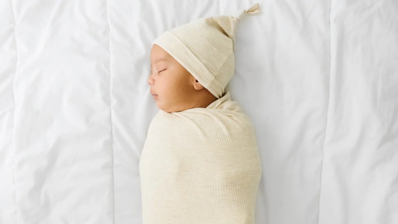 Swaddled baby peacefully sleeping on a white bed, wearing a cream-colored hat and wrap.