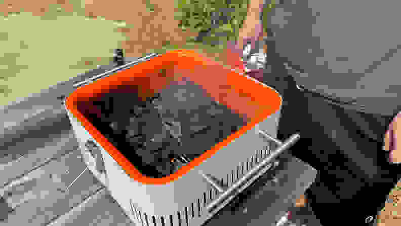 A person is standing beside an Everdure Cube grill in gray, which is packed with burning charcoal.