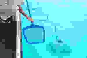 The image shows a pool vacuum cleaner working in the bottom of a pool, while someone uses a net.
