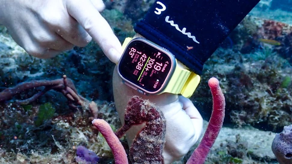 A gloved scuba diver wears a Apple Watch Ultra with the Oceanic+ app open as a seahorse floats in the foreground.