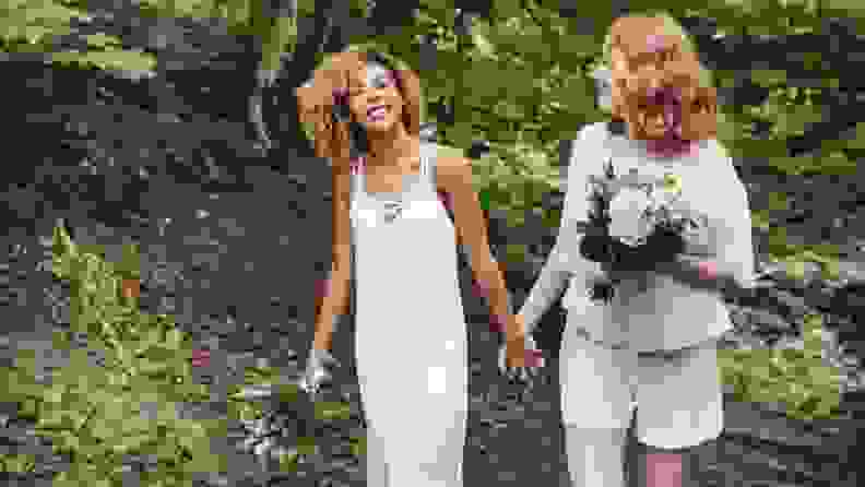 Two brides dressed in white outfits and carrying wedding bouquets walk through a wooded area