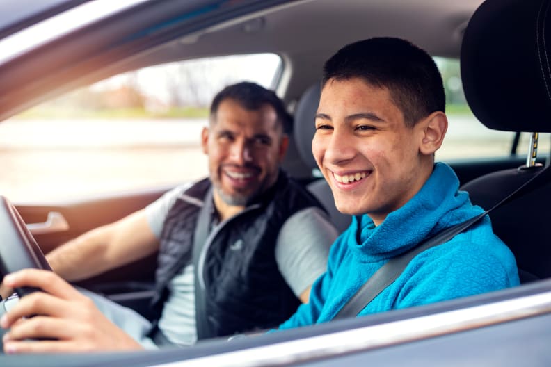 A teenager sits behind the wheel of a car and listens to his father's instructions as he drives.