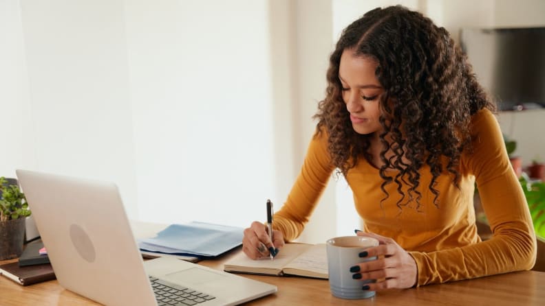 Woman writing in journal wearing mustard-colored shirt in front of laptop.