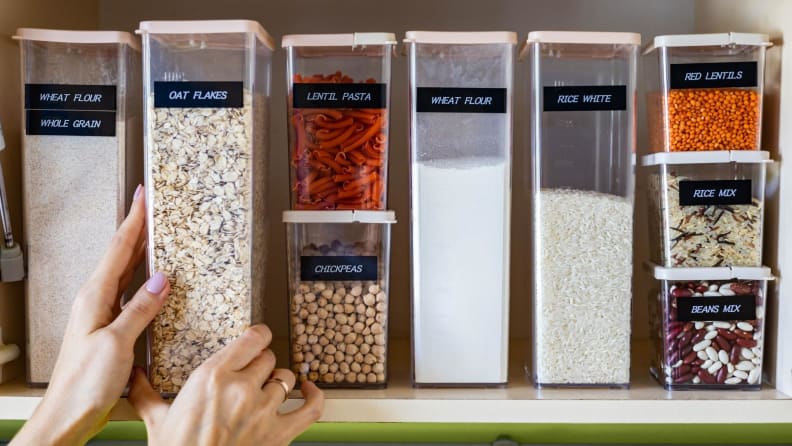 Food bins neatly organized and named in a pantry, with a hand reaching out for one of them.