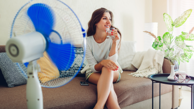 A blue fan spins while a woman sits on couch drinking water