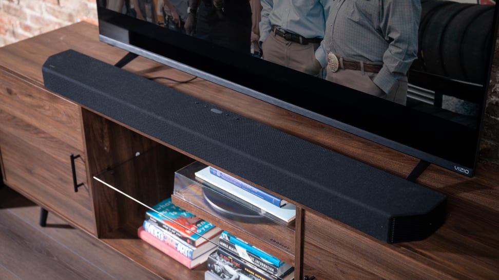A black soundbar sits on a walnut colored stand below a black TV and above some blu-rays.