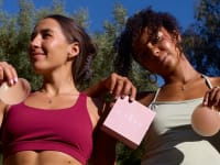 Two women wearing activewear hold a box and a pair of Cakes nipple covers outdoors in the sunlight.