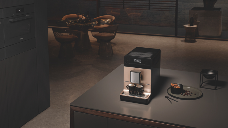 In a kitchen furnished with Miele appliances, a Miele CM5 coffee system with rose metallic finish is in the center making two shots of coffee. Some snacks are scattered around. There's a dining set in the background.