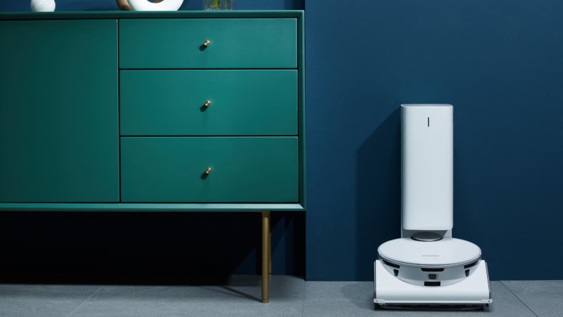 A white robot vacuum sits in its doc next to a green dresser