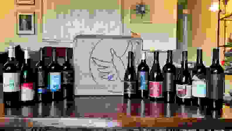 A row of wine bottles from Naked wine on top of wooden table surrounding a cardboard box.