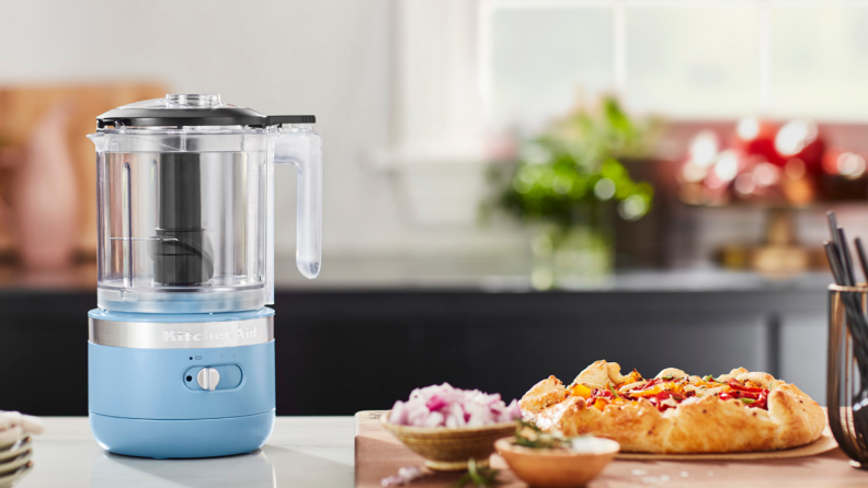 A blue KitchenAid 5-cup cordless mini food processor is seen on a kitchen counter, alongside some foods.