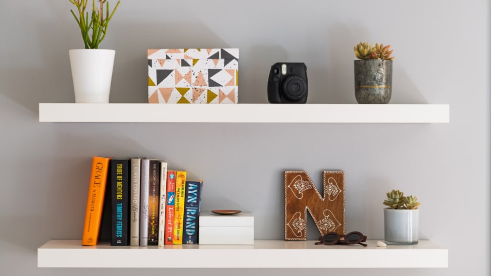 Decorations including a plant, jars, books, and boxes sit on two floating white shelves on a grey wall.