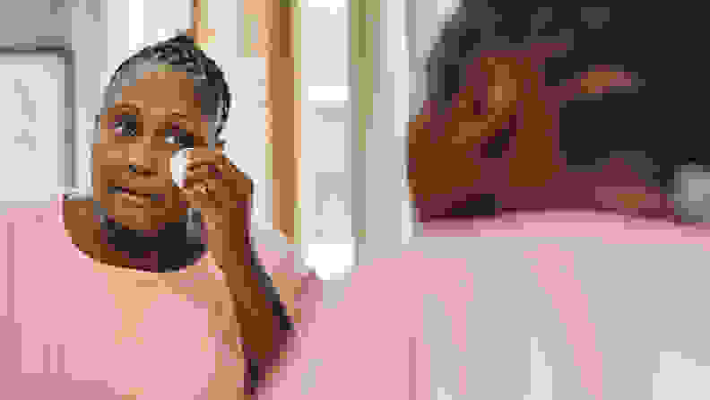 A person with dark skin looks into a mirror and removes eye makeup using a cotton round.