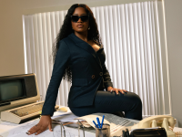 Actress Keke Palmer sits on a desk. She is wearing a black suit with dark sunglasses.