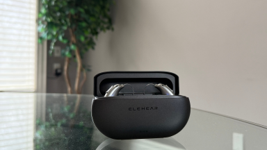 An open black charging case with hearing aids inside sits on top of a glass table with a tree in the background
