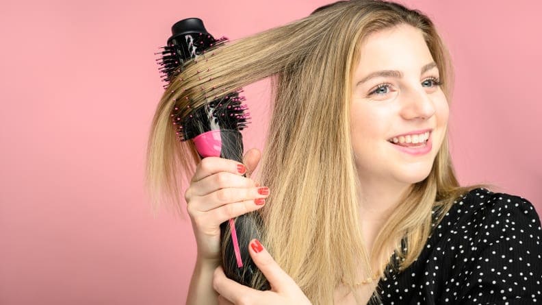 A blonde woman standing in front of a pink background styling her hair with a Revlon volumizer.