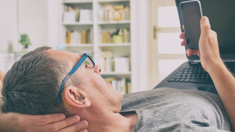 A person wearing a hearing aid lays in bed with a cell phone and laptop.