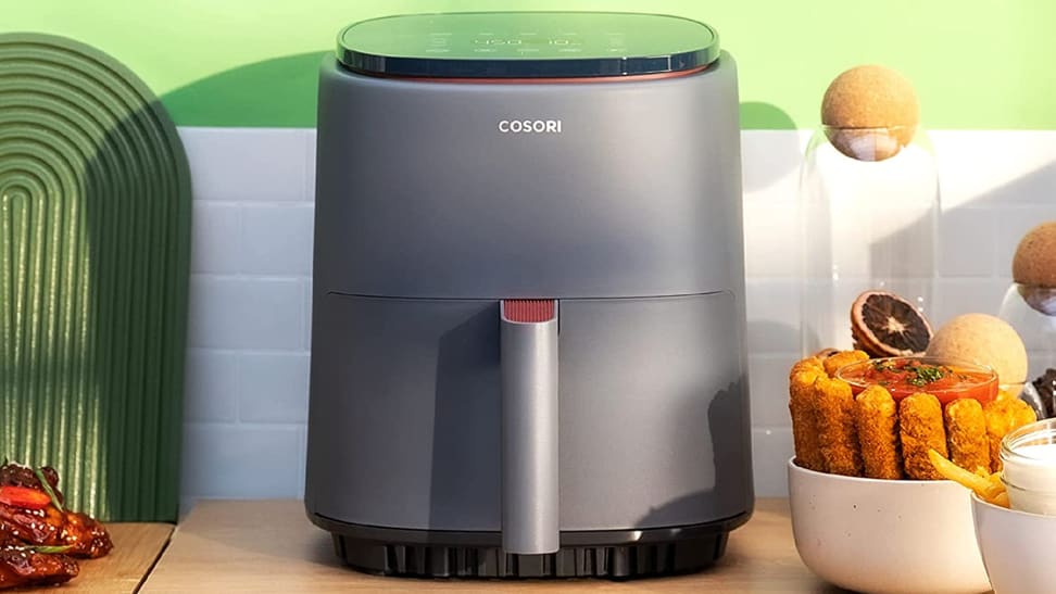 A Cosori Lite four-quart air fryer sitting on a kitchen counter in front of a green wall with white tiles and surrounded by food at home.