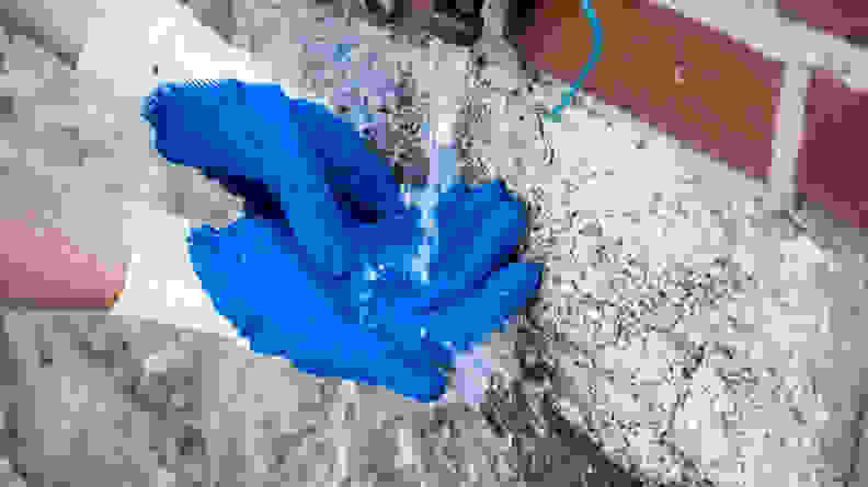 Two gardening-gloved hands are washing off dirt and grime using an outdoor water spigot.
