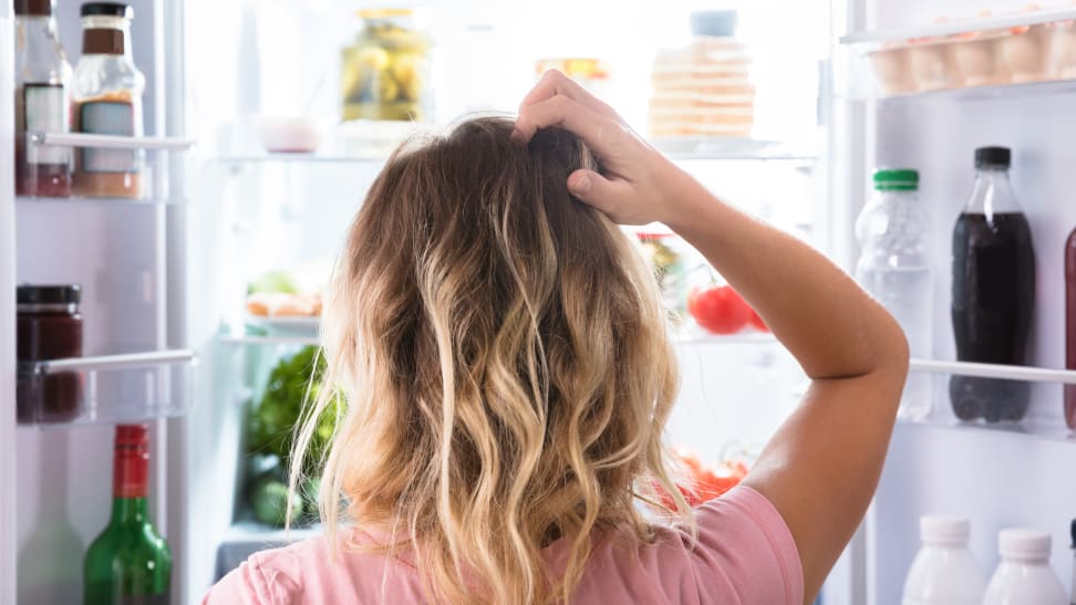 A woman stands in front of an open refrigerator, scratching her head.