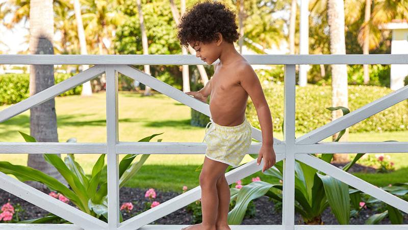 Little boy wearing swim trunks and standing on a railing