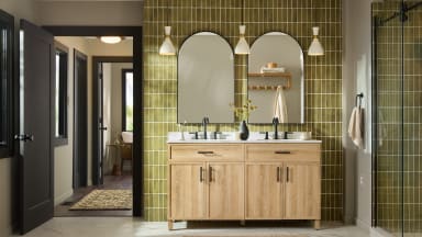 A modern bathroom vanity with a double sink sits against an olive-green tiled wall with two arched mirrors and brass-accent pendant lights.