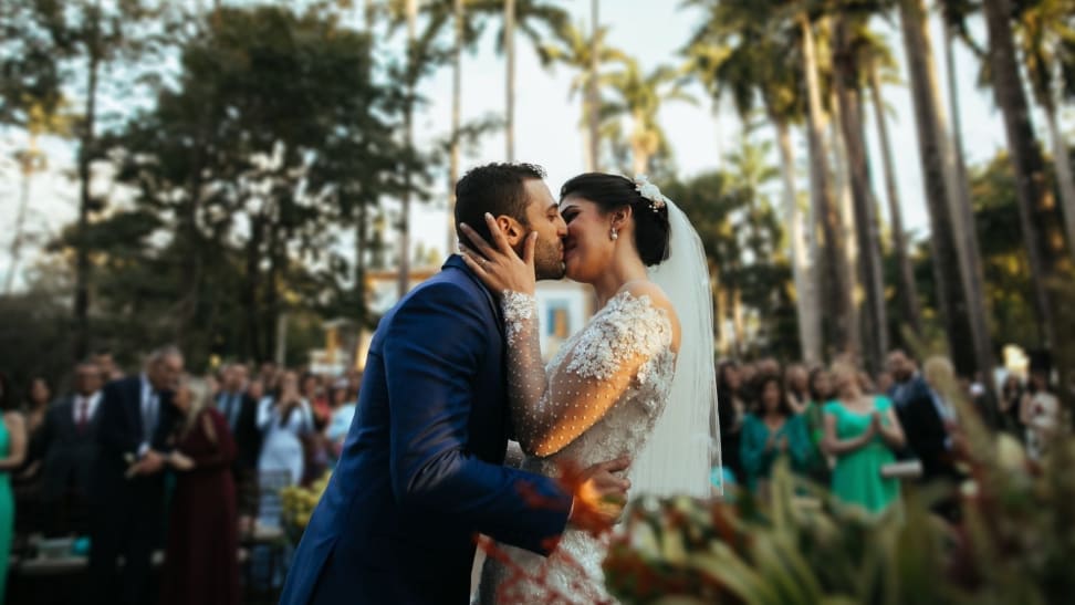 A bride and groom kiss in front of wedding guests with palm trees in the background