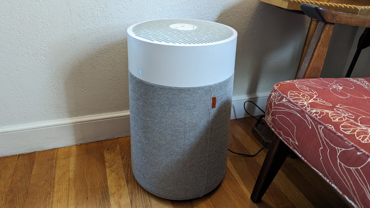 Gray and white Blueair Blue Pure 311i Max purifier sitting on top of wooden floor boards next to padded chair in front of white wall.