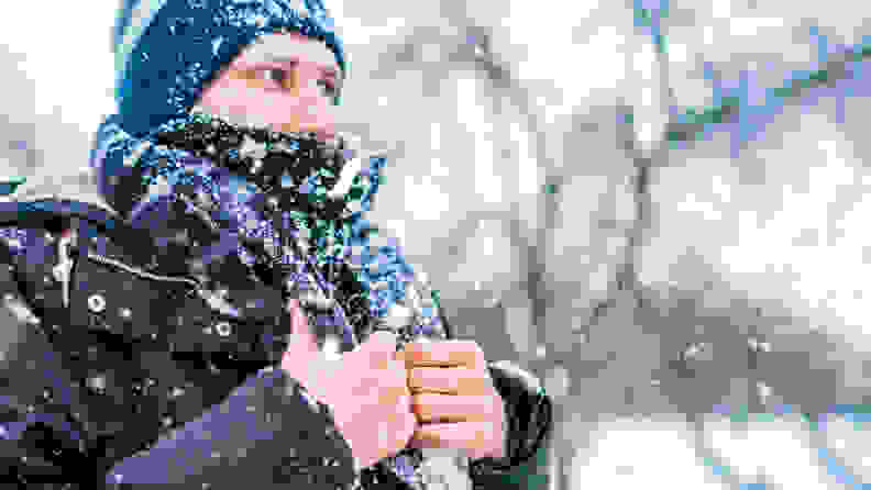 A person stands bundled up (sans gloves) while snow falls all around them.