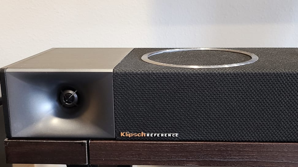 A silver and black soundbar is shown from the left side with a silver circlet above for the Dolby Atmos speaker, set on a brown console