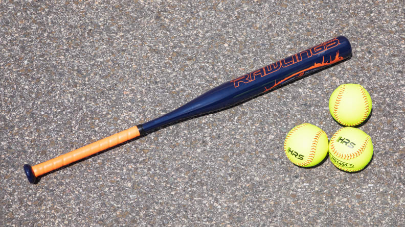 Three neon yellow softballs next to navy blue and orange baseball bat on carpeted floor.