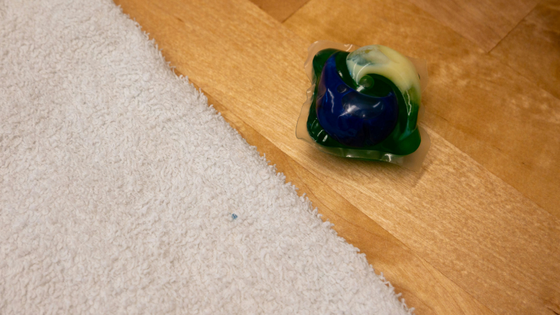 A detergent pod is shown next to a towel, with a blue detergent stain on it.