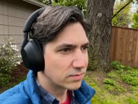 A man with brown and grey hair wears the matte black Sony WH-1000XM5 headphones in front of a large tree and a beige house.