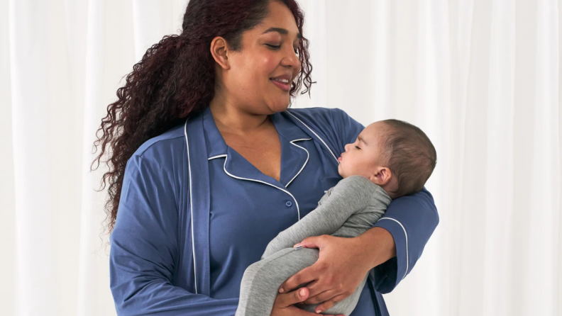 aregiver in blue pajamas smiling down at a sleeping baby dressed in a gray onesie.