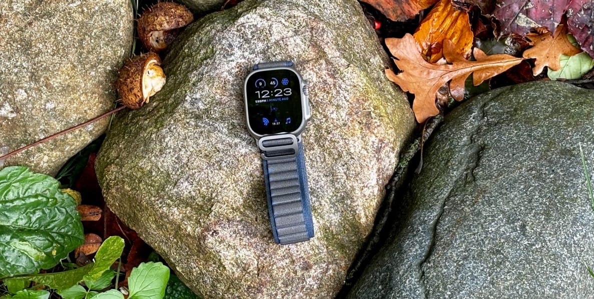 An Apple Watch Ultra 2, with a blue band, sits on a wet rock in the forest