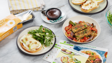 Four plates of HelloFresh meals, a package of egg bites, and recipe cards spread out on a counter.