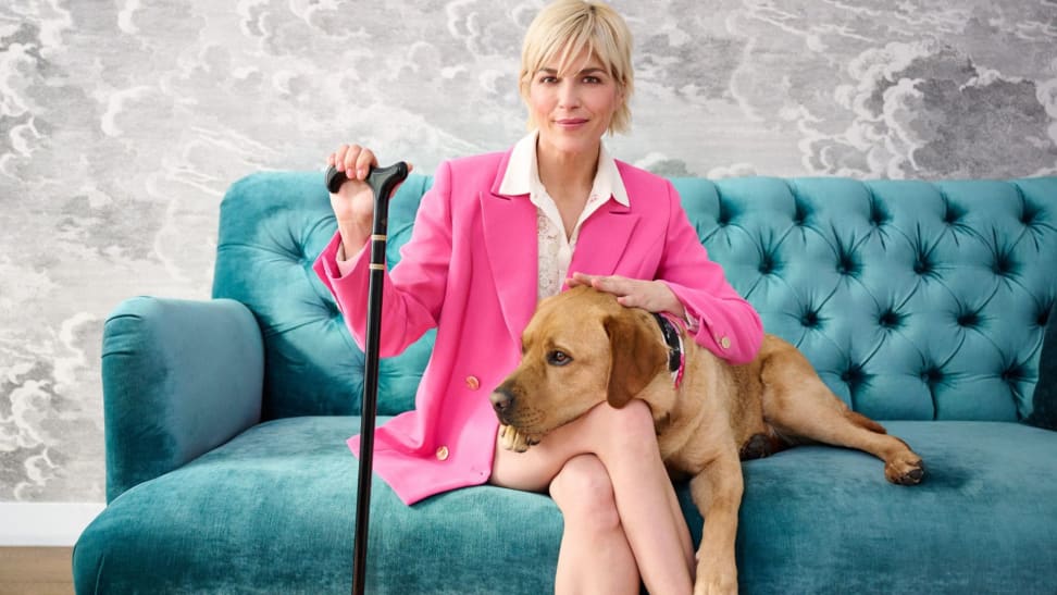 Selma Blair, QVC Brand Ambassador For Accessibility, Sits On A Plush Teal Sofa With Her Dog, Scout. She Wears A Hot Pink Suit And Uses A Black And Gold Cane.