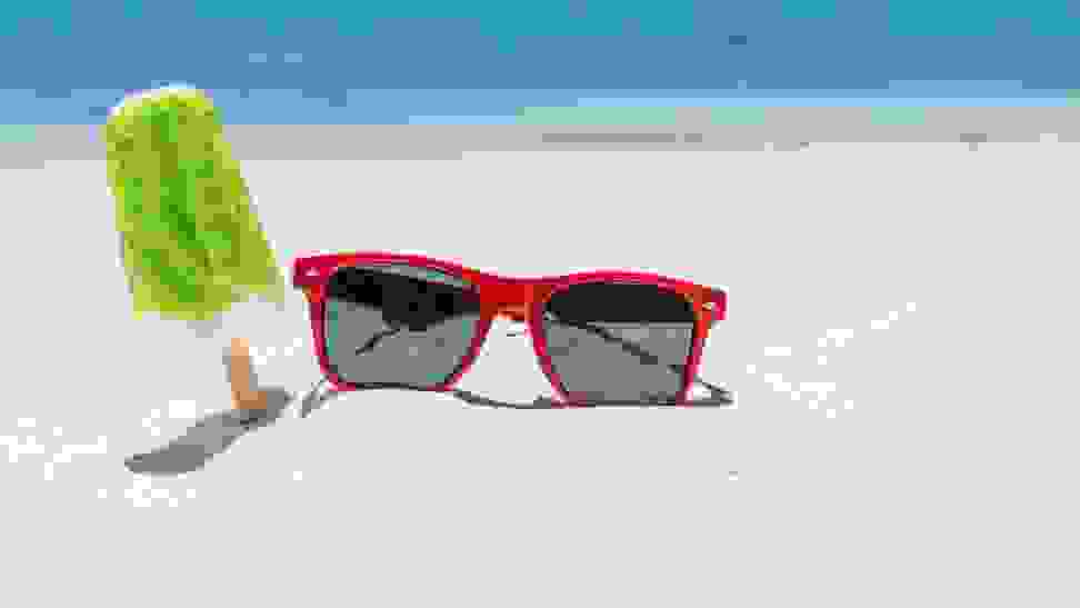 A pair of red Ray-Ban sunglasses lie on the sandy beach beside a green popsicle.