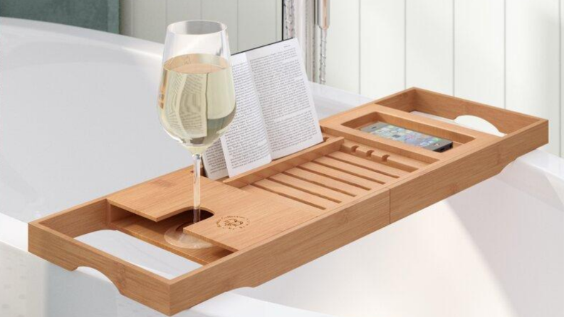 A wooden bath tray holding a wine glass, a book, and a cell phone over the bathtub