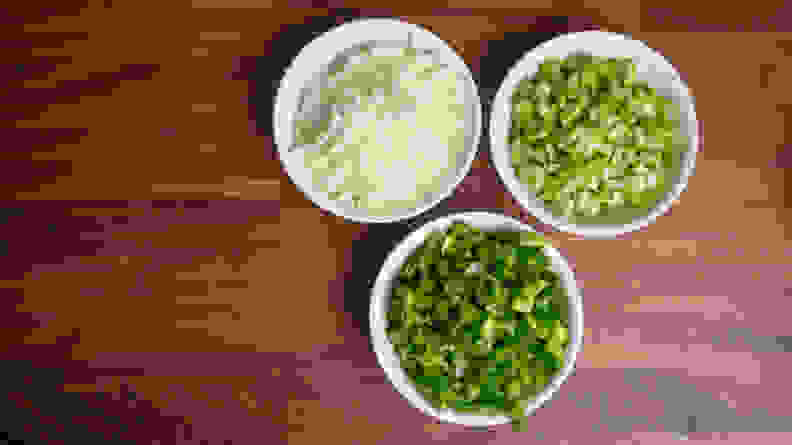 Three small white bowls of bell pepper, onion, and celery arranged together on a countertop, shot from above.